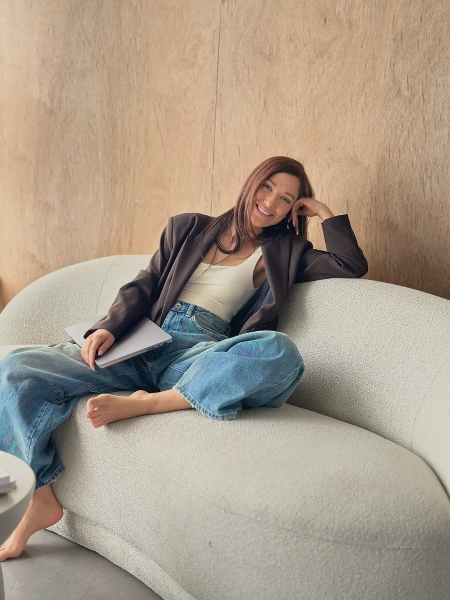 Diyana Bancheva sitting on a light textured sofa with a laptop, wearing a dark blazer and jeans, smiling at the camera against a wood-panelled wall