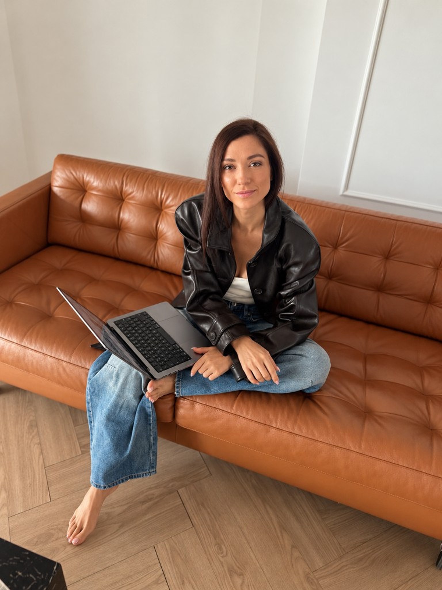 Diyana Bancheva sitting cross-legged on a tan leather sofa, wearing a black leather jacket and wide-leg jeans, working on a laptop